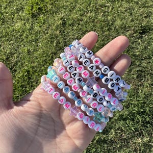 Könnte beinhalten: Eine Sammlung von Armbändern mit Perlen, die "TEAM BOY" und "TEAM GIRL" mit weißen Buchstabenperlen buchstabieren. Die Armbänder sind in verschiedenen Farben gehalten, darunter Rosa, Blau und Transparent, mit sternförmigen und runden Perlen. Die Armbänder werden in einer Hand vor einem Hintergrund aus grünem Gras gehalten.