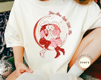 May include: Ivory-colored t-shirt with a graphic of two cowgirls on a crescent moon, with the text "You Are Safe With Me." The design is in shades of red. The shirt is labeled "Comfort Colors" and "Ivory."