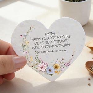 Puede incluir: Una tarjeta en forma de corazón con detalles florales y el texto "Mamá, gracias por criarme para ser una mujer fuerte e independiente". También se ven una pequeña maceta blanca con romero y lavanda, una cuchara de madera y una pequeña bolsa de semillas.