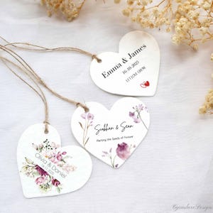 Puede incluir: Etiquetas de recuerdo de boda en forma de corazón, blancas, con diseños florales y nombres. Las etiquetas tienen hilo marrón y presentan nombres, fechas y frases como "Let Love Grow" y "Planting the Seeds of Forever".