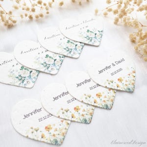 Puede incluir: Tarjetas de mesa de boda en forma de corazón con diseños florales y nombres. Algunas tarjetas presentan nombres como "Jennifer" y "David" con fechas. Otras tienen nombres como "Karstine" y "Alexander" con fechas. Las tarjetas están decoradas con flores de acuarela.