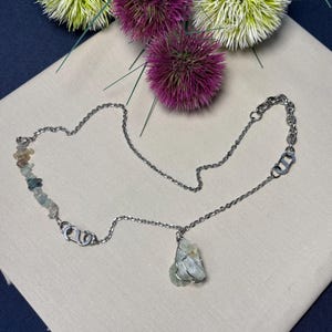May include: A silver necklace with a raw, pale blue gemstone pendant. The chain has small, colourful stone accents and decorative links. The jewellery is displayed on a neutral fabric with purple and yellow floral accents.