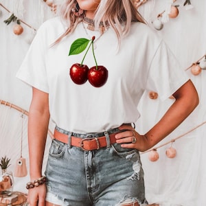 May include: White t-shirt featuring a vibrant graphic of two ripe cherries with a green leaf. The cherries are a deep red color with water droplets, creating a realistic look. The shirt is paired with denim shorts and a brown belt.