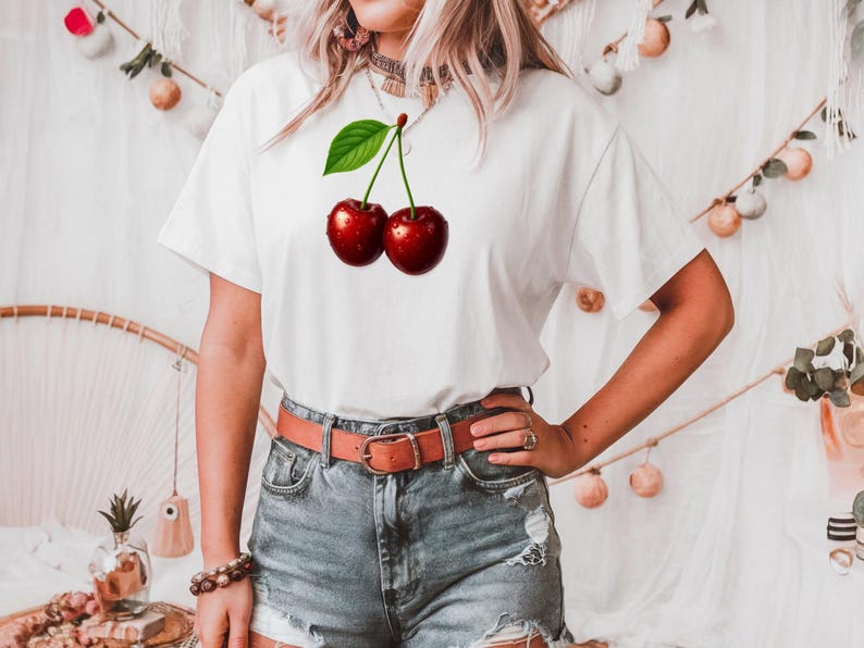May include: White t-shirt featuring a vibrant graphic of two ripe cherries with a green leaf. The cherries are a deep red color with water droplets, creating a realistic look. The shirt is paired with denim shorts and a brown belt.
