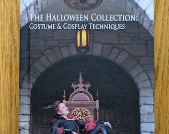 May include: Book cover featuring a woman in a red and black costume sitting on a throne with a whip in her hand. The title of the book is "The Halloween Collection: Costume & Cosplay Techniques" by Laura Meyer.