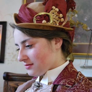 May include: A vintage-style hat with a burgundy and tan color scheme. The hat features a decorative gold buckle and trim, with a matching burgundy ribbon and feather accents. The hat is part of a historical costume.