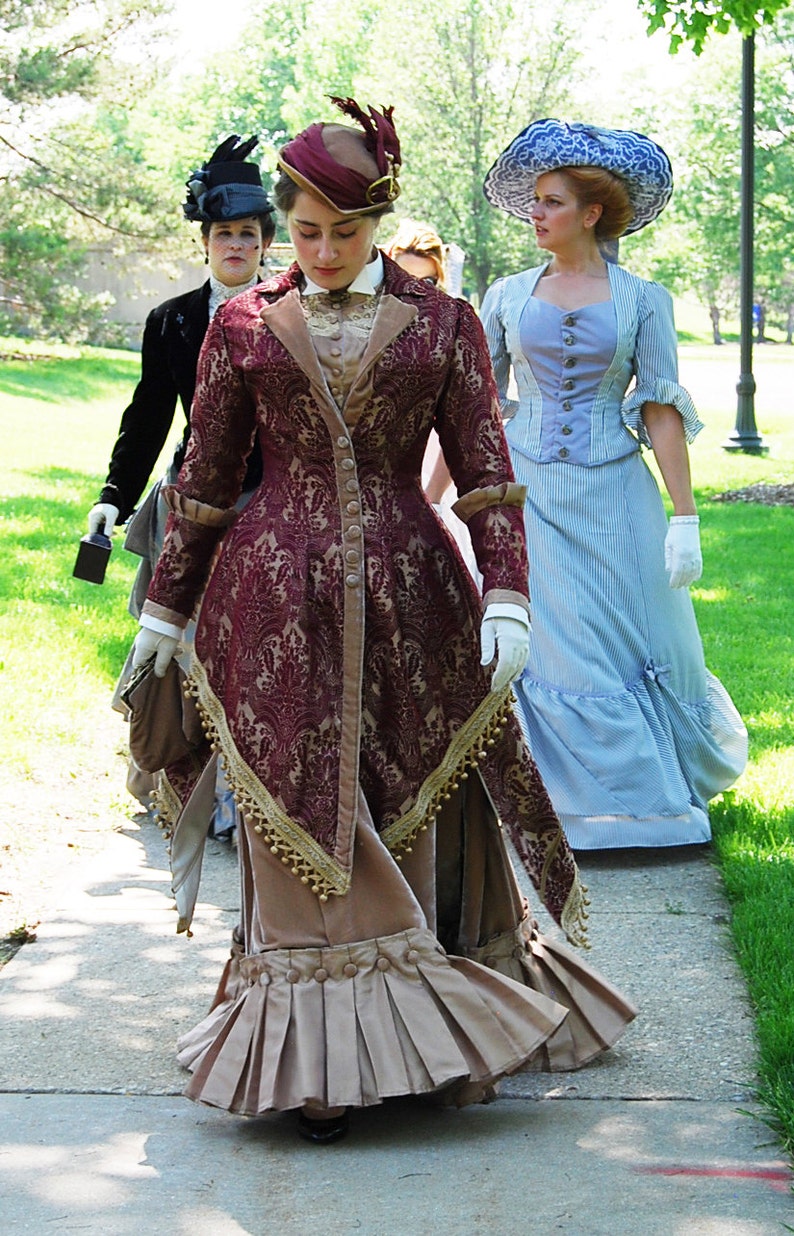 May include: A group of people in vintage Victorian-era dresses walking on a path. The dresses are in various colors, including burgundy, tan, and blue. The burgundy dress has a detailed floral pattern and gold trim. The women are wearing hats and gloves.