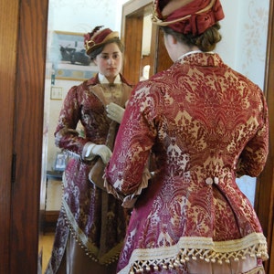 May include: A woman in a burgundy and gold patterned Victorian era dress and matching hat, reflected in a mirror. The dress features intricate detailing, including buttons, trim, and a layered skirt. The hat is adorned with a feather.