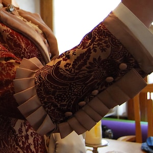 May include: Close-up of a sleeve from a burgundy and gold patterned jacket. The sleeve features a decorative cuff with pleats and small, round buttons. The jacket appears to be part of a historical costume or formal wear.