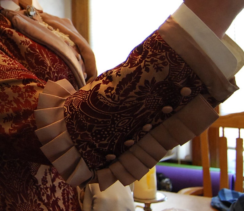 May include: Close-up of a sleeve from a burgundy and gold patterned jacket. The sleeve features a decorative cuff with pleats and small, round buttons. The jacket appears to be part of a historical costume or formal wear.