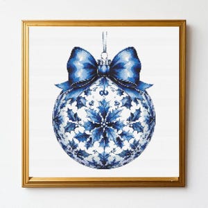 Könnte beinhalten: Gerahmter, kreuzgestickter Weihnachtsschmuck. Der Schmuck ist eine blau-weiße Kugel mit einem Stechpalmenblattmuster und einer blauen Schleife oben. Der Rahmen ist goldfarben.