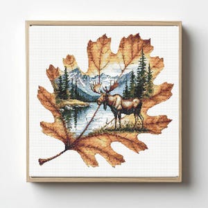 May include: Cross-stitch art of a moose standing near a lake, framed within a large, detailed autumn leaf. The leaf is a mix of brown and orange hues, with a light-colored background. The scene includes mountains and evergreen trees.