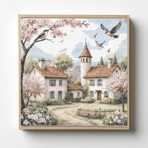 May include: A framed cross-stitch landscape depicts a quaint village scene. The artwork features houses with red tile roofs, a tower, and blossoming trees in shades of pink. Birds fly in the sky above the buildings. The artwork is framed in a light wood.