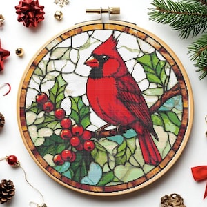 May include: A cross-stitch embroidery in a wooden hoop features a vibrant red cardinal perched on a branch with holly berries. The design resembles stained glass, with green leaves and a white background. Christmas-themed decorations surround the hoop.