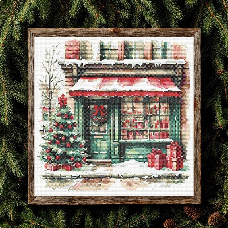 May include: A cross-stitch pattern depicting a storefront with a red awning, a decorated Christmas tree, and wrapped gifts, all framed in wood. The scene features snow-covered surfaces, a green door with a wreath, and a window display of items.