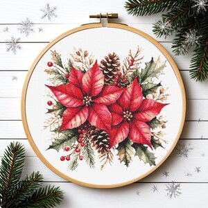 Op de afbeelding: Kruissteekborduurwerk van een poinsettia bloemstuk in een houten hoepel. Het ontwerp toont grote rode poinsettia bloemen, groene hulstbladeren, dennenappels en rode bessen, perfect voor de feestdagen.