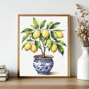 May include: Cross-stitch artwork of a lemon tree in a blue and white patterned pot, set in a wooden frame. The tree has green leaves and several yellow lemons. The artwork is displayed on a wooden surface.