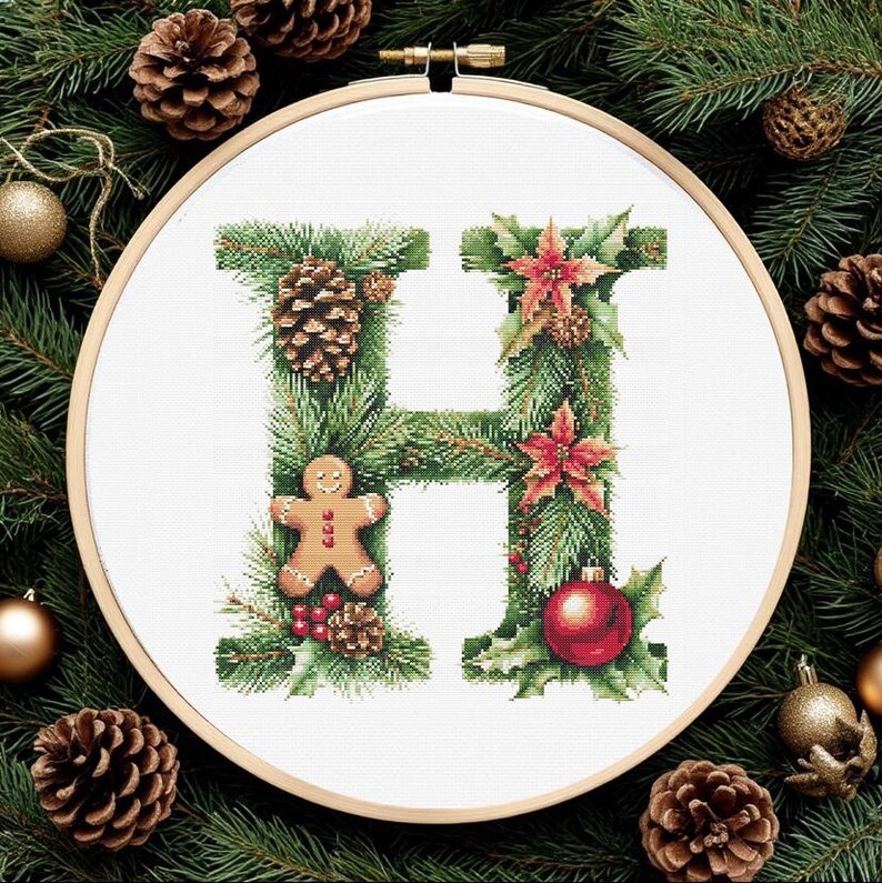 May include: Cross-stitch embroidery of the letter "H" decorated with Christmas elements. The design includes pine needles, pine cones, red poinsettias, a gingerbread man, and a red ornament, all set within a wooden embroidery hoop.