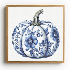May include: A square framed cross-stitch artwork featuring a white pumpkin decorated with intricate blue floral patterns and leaves. The pumpkin has a textured green and brown stem and is centered on a white fabric background within a light wood frame.