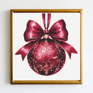 Puede incluir: Un bordado a punto de cruz enmarcado de un adorno navideño. El adorno es de color rojo intenso con detalles florales dorados y un lazo a juego. El marco es de color dorado cálido y el fondo es blanco.