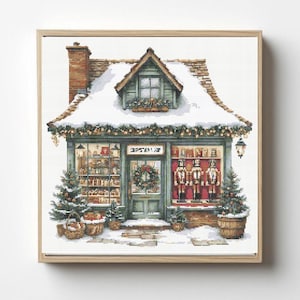 May include: A cross-stitch pattern depicting a snow-covered shop with a brick chimney, a green storefront, a central door with a wreath, and display windows showing shelves of items and three nutcracker figures, framed in light wood.
