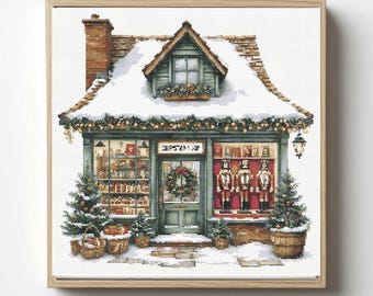 May include: A cross-stitch pattern depicting a snow-covered shop with a brick chimney, a green storefront, a central door with a wreath, and display windows showing shelves of items and three nutcracker figures, framed in light wood.