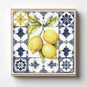 May include: A square cross-stitch embroidery piece featuring three bright yellow lemons with green leaves centered against a background of blue and white decorative tile patterns, all enclosed within a light-colored wooden frame.