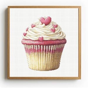May include: Cross-stitch art of a cupcake with white icing and pink heart sprinkles. The cupcake has a pink base and a yellow and white striped wrapper. The artwork is framed in a light brown wooden frame.
