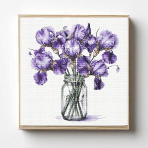May include: A framed cross-stitch of a bouquet of purple irises in a clear glass jar. The irises have ruffled petals and are arranged in a full, lush display. The jar is clear, and the background is white.