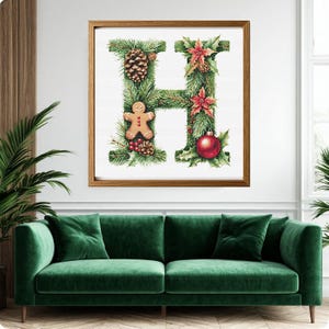 May include: A framed Christmas-themed artwork featuring the letter "H" decorated with pine needles, pine cones, red berries, a gingerbread man, poinsettias, and a red ornament. The artwork is displayed above a green velvet sofa.