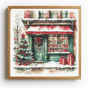 May include: A square cross-stitch embroidery in a light wood frame depicting a storefront with a red awning, a green door with a wreath, and a large Christmas tree decorated with red ornaments, all covered in snow with red gift boxes.