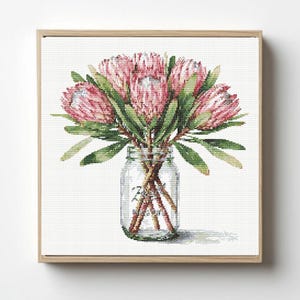 May include: A cross-stitch embroidery pattern featuring a bouquet of pink protea flowers with green leaves arranged in a clear glass mason jar. The design is framed in a light-colored wooden square frame, showcasing detailed needlework stitches.
