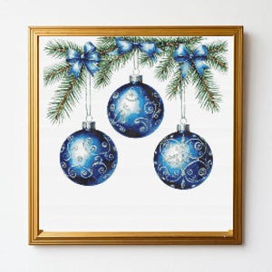 May include: A framed cross-stitch embroidery featuring three round blue Christmas baubles with silver swirl patterns, hanging from green pine branches decorated with three blue bows. The artwork is enclosed in a simple gold-coloured frame.