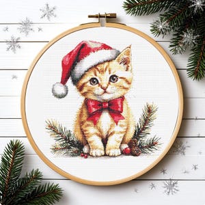 May include: A cross-stitch embroidery project in a round wooden hoop featuring a ginger tabby kitten wearing a red and white Santa hat and a red bow tie, surrounded by green pine branches and small red berries on a white fabric background.
