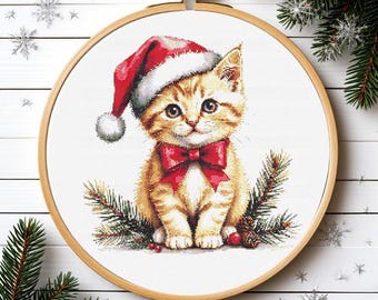May include: A cross-stitch embroidery project in a round wooden hoop featuring a ginger tabby kitten wearing a red and white Santa hat and a red bow tie, surrounded by green pine branches and small red berries on a white fabric background.