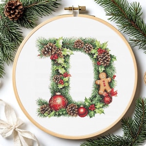 May include: Cross-stitch embroidery of the letter "D" in a wooden hoop, decorated with Christmas greenery, pinecones, red berries, and ornaments. A gingerbread man is also included in the design. The embroidery is set against a white background.