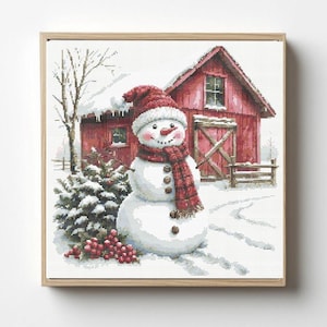 May include: A cross-stitch pattern featuring a snowman wearing a red knit hat and scarf, standing in a snowy landscape with a red barn, a small evergreen tree, and red berries, all framed in a light-colored wooden square frame.