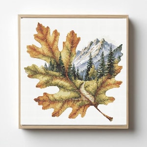 Può includere: Opera d'arte a punto croce incorniciata raffigurante una foglia autunnale con una scena di montagna e foresta. La foglia è nei toni del marrone e del verde, con un sentiero che si snoda tra gli alberi. La cornice è in legno chiaro.