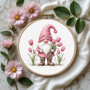 May include: A cross-stitch embroidery in a wooden hoop features a pink gnome holding tulips. The gnome wears a pink hat and coat, with a white beard. Pink tulips and flowers surround the gnome, with additional flowers in the background.