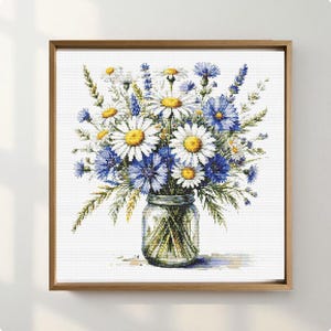 May include: A framed cross-stitch of a floral arrangement in a glass jar. The bouquet features white daisies with yellow centers, blue cornflowers, and green foliage. The artwork is set against a white background and framed in a light brown wooden frame.
