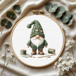 Könnte beinhalten: Ein Kreuzstich-Stickrahmen mit einem Zwerg mit grünem Hut und Pullover, weißem Bart und braunen Stiefeln. Der Zwerg ist umgeben von Garnrollen, einem Lineal und Zweigen von Eukalyptus und weißen Blumen.