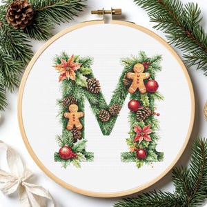 Könnte beinhalten: Kreuzstich-Stickerei des Buchstabens "M", verziert mit Weihnachtselementen. Der Buchstabe ist mit grünen Tannennadeln, Tannenzapfen, roten Kugeln, Weihnachtssternen und Lebkuchenmännern geschmückt, alles in einem hölzernen Stickrahmen.