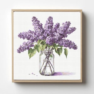 Peut inclure: Une broderie au point de croix encadrée de lilas violets dans un bocal en verre transparent. Les fleurs sont en pleine floraison avec des feuilles vertes. Le bocal est posé sur un fond blanc, et le cadre est en bois clair.