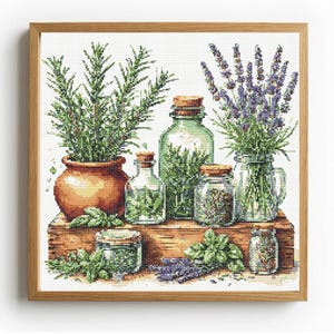 Op de afbeelding: Een kruissteekpatroon met een rozemarijnplant in een terracotta pot, lavendel in een glazen pot en diverse glazen potten gevuld met kruiden en specerijen, gerangschikt op een houten ondergrond in een lichthouten lijst.