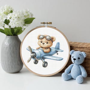 Può includere: Un telaio da ricamo circolare in legno contenente un motivo a punto croce di un orsacchiotto marrone chiaro con occhiali da aviatore che pilota un aereo a elica azzurro. Il disegno è centrato su uno sfondo in tessuto bianco.