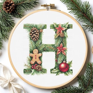 May include: Cross-stitch embroidery of the letter "H" decorated with Christmas elements. The design includes pine needles, a pine cone, a gingerbread man, red poinsettias, and a red ornament. The embroidery is set in a wooden hoop.