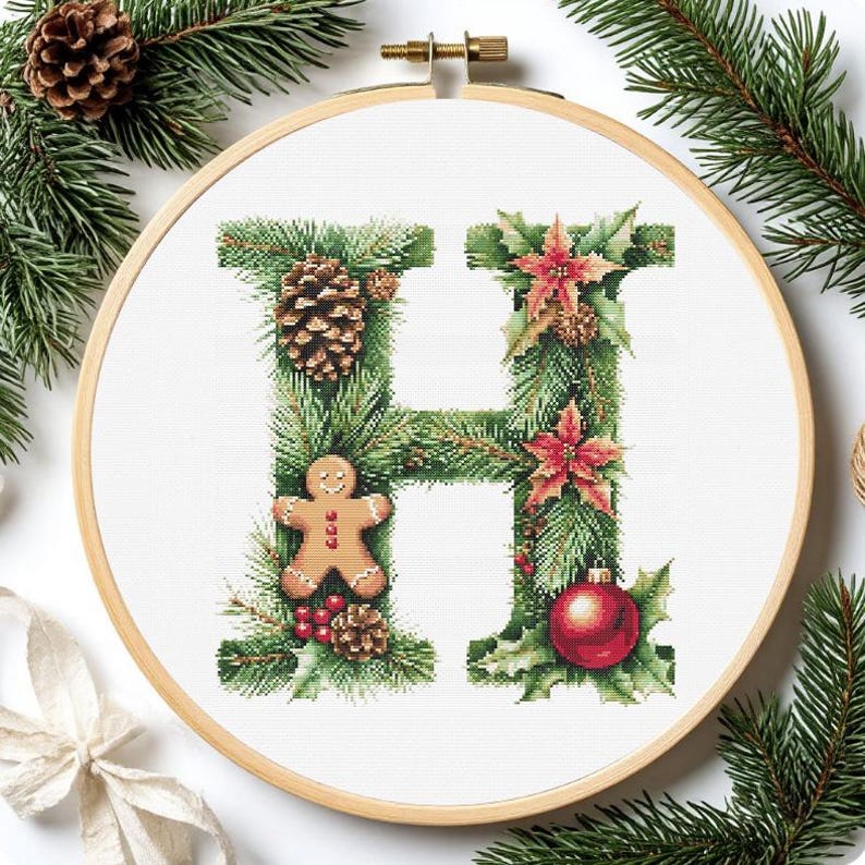 May include: Cross-stitch embroidery of the letter "H" decorated with Christmas elements. The design includes pine needles, a pine cone, a gingerbread man, red poinsettias, and a red ornament. The embroidery is set in a wooden hoop.
