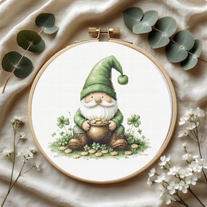 May include: A cross-stitch embroidery hoop featuring a whimsical gnome design. The gnome, dressed in green, holds a pot of gold amidst shamrocks. The embroidery is set in a wooden hoop, with sprigs of greenery and white flowers around the edge.