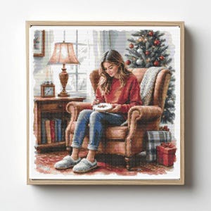 Puede incluir: Una impresión enmarcada representa a una persona con un suéter rojo y jeans azules sentada en un sillón marrón, sosteniendo un plato. Un árbol de Navidad, regalos y una lámpara son visibles en el fondo. La obra tiene una estética cálida.