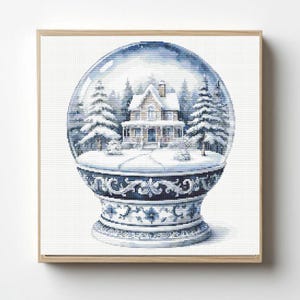 May include: A square framed cross-stitch embroidery depicting a snow globe containing a two-story house surrounded by snow-covered pine trees. The base of the globe features intricate blue and white floral patterns on a dark blue background.